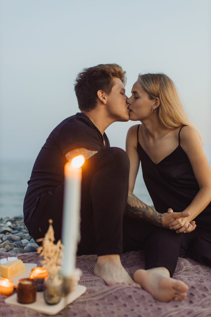 A couple shares a tender kiss during a romantic beach picnic surrounded by candlelight.