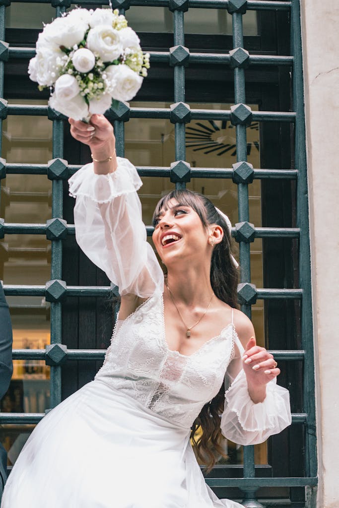 A happy bride in a white dress joyfully throws her wedding bouquet outdoors, celebrating love and marriage.