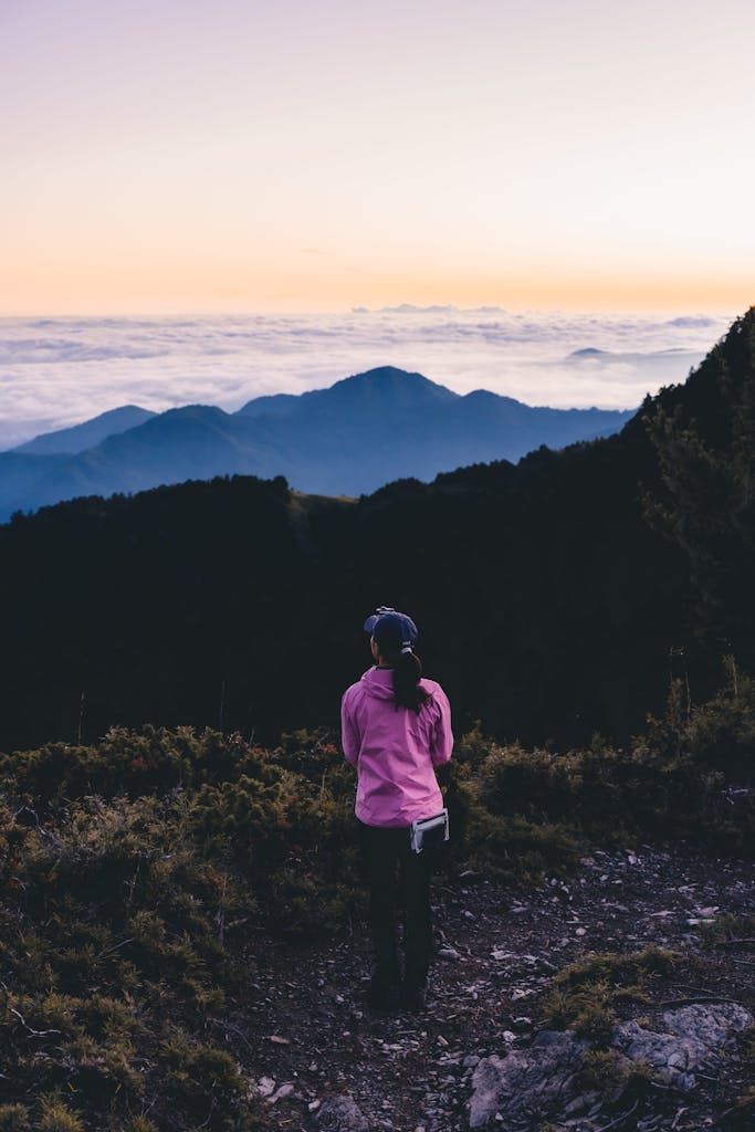 A woman in a pink jacket stands on a mountain path overlooking a cloud-filled valley at sunrise.