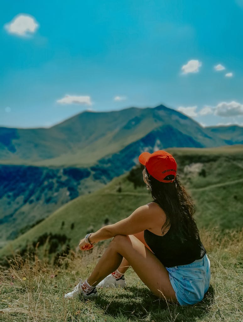 A woman sits on a grassy hillside wearing a red cap, enjoying a view of mountains under a clear blue sky.