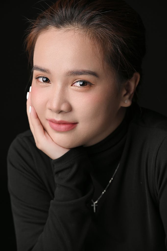 Close-up studio portrait of an Asian woman with hand on cheek against a black background.