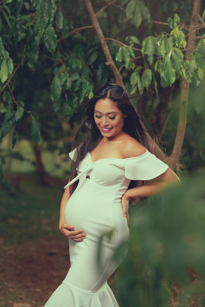 Pregnant woman in a white dress, posing outdoors amidst lush greenery, conveying joy and anticipation.