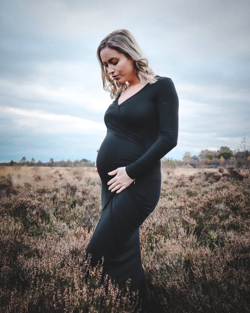 Pregnant woman in black dress posing outdoors in a tranquil field setting.