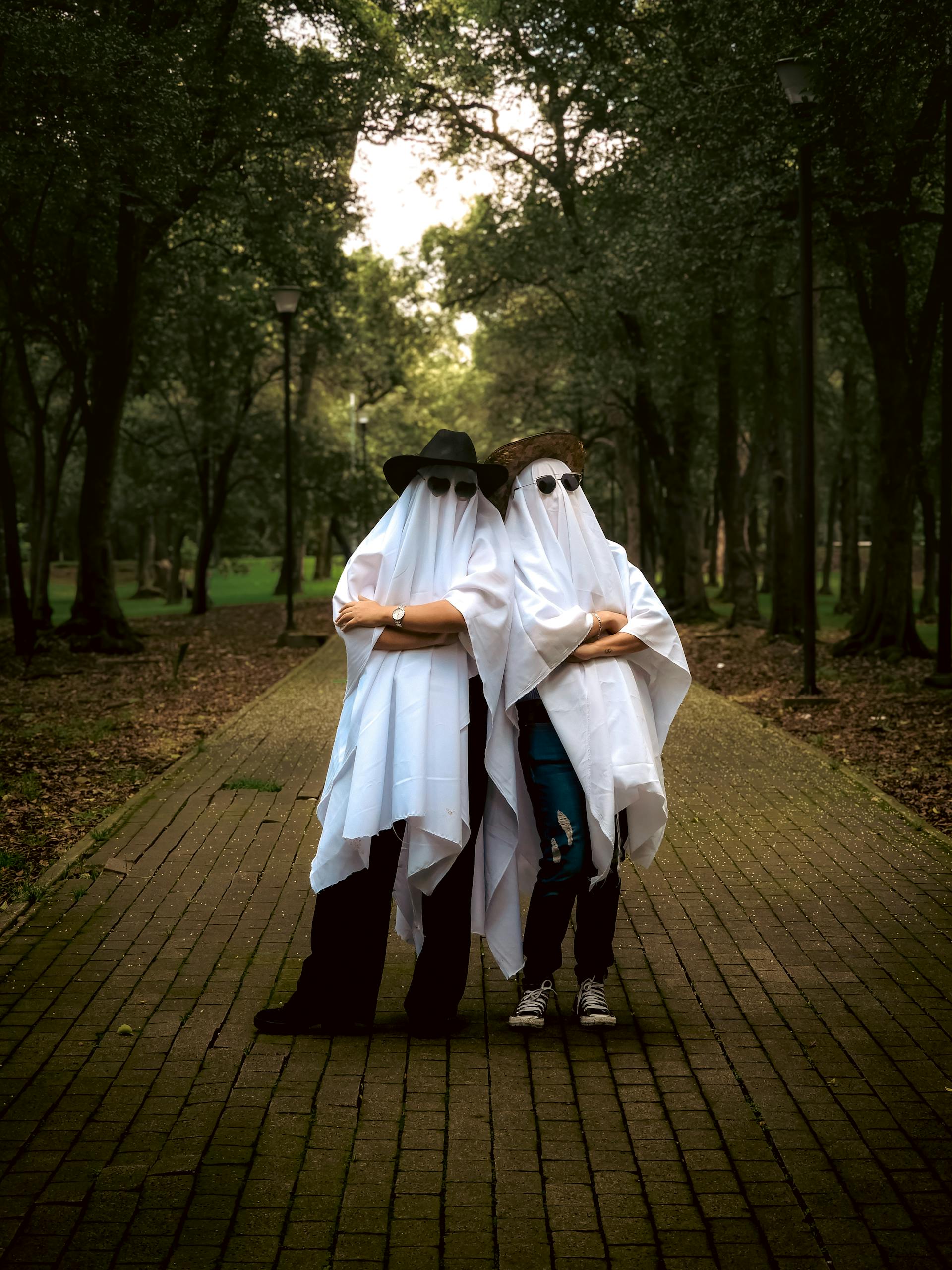 Two people in ghost costumes pose playfully in a wooded park path.